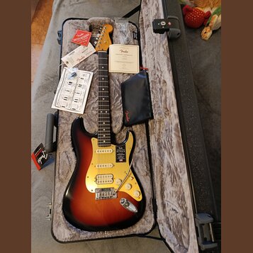 Elektrická kytara typu Stratocaster s barevným přechodem od černé přes červenou k žluté a světlým hmatníkem. Na krytce je nápis „American ULTRA STRAT…