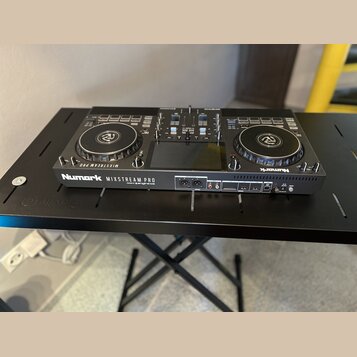 Černý DJ 컨트롤러 Numark MIXSTREAM PRO umístěný na černém stole. Na přední straně je vidět bílý nápis Numark MIXSTREAM PRO a menší text pod ním, který je…