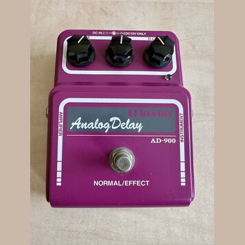 Fialový efekt pedál s nápisem Maxon Analog Delay a označením modelu AD-900. Na horní straně jsou tři černé oválné knoflíky a nápis DC IN.(-)—(+)DC12V…