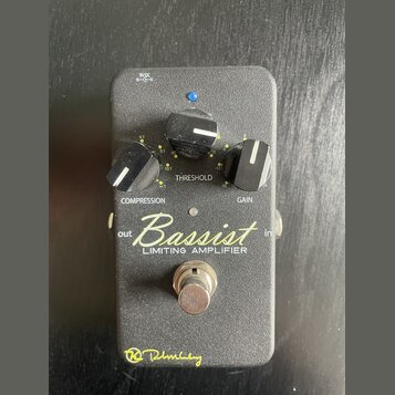 Černý kytarový pedál s nápisem Bassist LIMITING AMPLIFIER a žlutým podpisem Rlhmberg ve spodní části. Obsahuje tři černé ovládací knoflíky s popisky…
