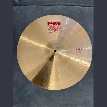Zlatavý kovový činel s viditelným broušením. Na povrchu je červené logo Paiste 2002 s nápisy TRADE MARK a MADE IN SWITZERLAND. Na pravé straně je čer…