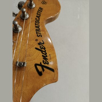 Detail hlavy kytary z dřeva s lakovaným povrchem. Na hlavě je černý nápis Fender® STRATOCASTER a malé vertikální sériové číslo 3143028. Jsou vidět ko…