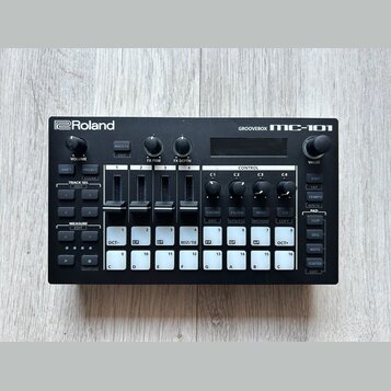 Černá groovebox s nápisy Roland a GROOVEBOX MC-101. Zařízení obsahuje displej, několik otočných ovladačů a bílá tlačítka s různými označením, včetně…