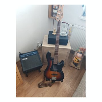 Elektrická basová kytara s tmavě hnědým sunburst povrchem a hmatníkem z tmavého dřeva. Na hlavě nástroje je bílý nápis Cort. Vedle kytary stojí malý…