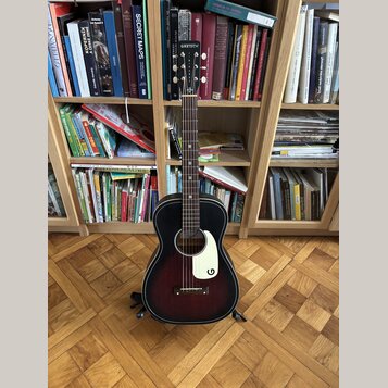 Akustická kytara s tmavě červeným gradientem (sunburst) a bílým plektrem. Na hlavě hmatníku je bílý nápis GRETSCH a na plektru bílé logo G. Nástroj j…