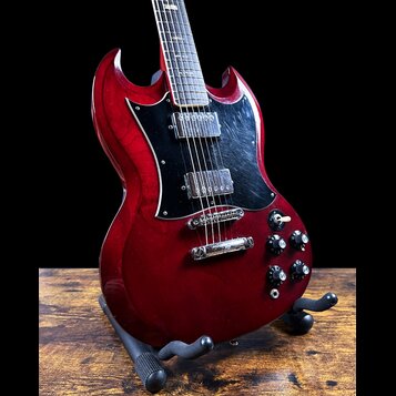 Elektrická kytara s červeným lakem a černým pickguardem, umístěná na černém stojanu. Nástroj má dva humbuckery a čtyři ovládací knoflíky. Povrch laku…