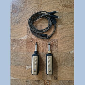 Na fotografii jsou dva zlatavé kable s černými plastovými koncovkami a jeden samostatný pletený kabel v hnědo-černé barvě. Zlatavé kable mají na povr…