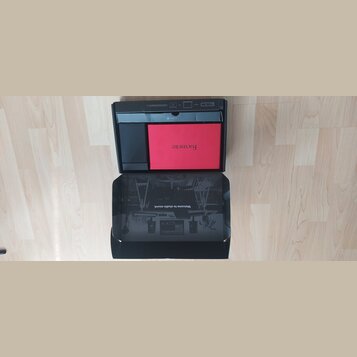 Fotografie znázorňuje otevřenou černou krabici s příslušenstvím. Uvnitř je červený čtvercový předmět s nápisem Focusrite. Na vnitřní straně víka je č…
