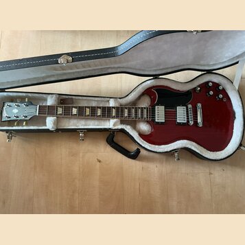 V otevřeném černém kufru s bílou vnitřní výplní leží červená elektrická kytara. Na hlavě nástroje je viditelné logo Gibson. Kytara má dva humbuckery,…