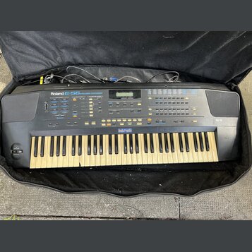 Černý digitální syntetizátor v černé přepravní brašně. Na přístroji je bílý nápis Roland E-56 SYNTHESIZER. Klávesnice vykazuje zežlutnutí bílých kláv…