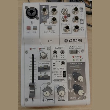 Bílá míchací konzole s nápisem YAMAHA a označením AG03 MIXING CONSOLE. Horní část obsahuje XLR vstup, dva jacky 6,35 mm s nápisy 2G GUITAR a LINE, or…