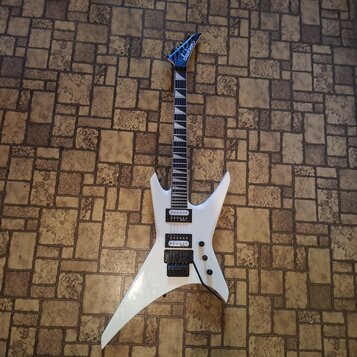 Bílá elektrická kytara s ostrými hranami korpusu a šicovým hmatníkem. Na hlavě je modré logo Jackson. Nástroj má dva humbuckery, jeden potenciometr a…
