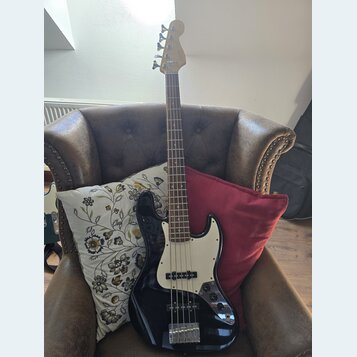 Černá elektrická basová kytara s bílým pickguardem a světlou hnedou hmatníkem. Na hlavě nástroje je viditelné logo Fender. Kytara je umístěna v hnědé…