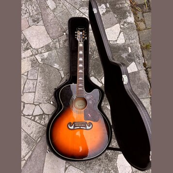 Akustická kytara s korpusem v barvě sunburst, umístěná v černém tvrdém pouzdře. Na hlavě hředu je bílý nápis Epiphone a na úzkém pásku v místě mostu…