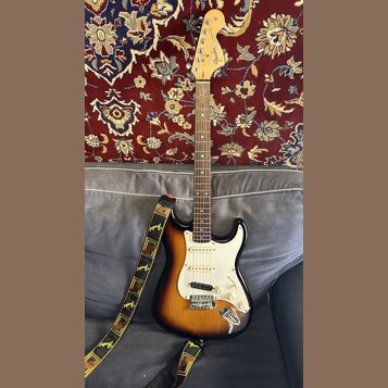 Elektrická kytara typu Stratocaster s barevným přechodem těla od žluté přes oranžovou k hnědé. Na hlavě hmatníku je viditelné logo Fender. Kytara má…