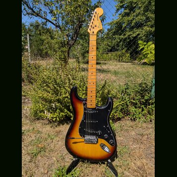 Elektrická kytara s tónem těla v přechodové barvě od černé po žlutou a černým pickguardem. Na hlavě hředu je vidět nápis Squier STRATOCASTER. Kytara…