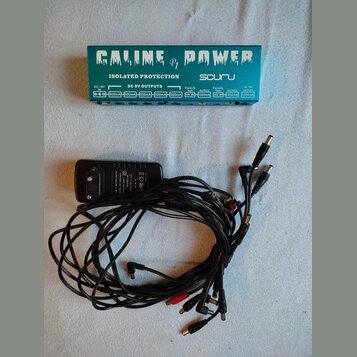Zelený napájecí adaptér pro pedály s nápisy GALINE P1 POWER a ISOLATED PROTECTION. Na pravé straně je logo souru a pod ním řada výstupů s označením D…