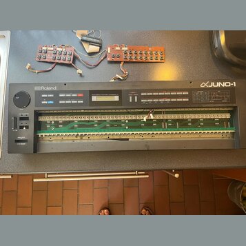 Šedý syntetizátor s nápisy Roland a JUNO-1, který je otevřený a vnitřní elektronika s deskami plošných spojů je viditelná. Na ovládacím panelu je náp…