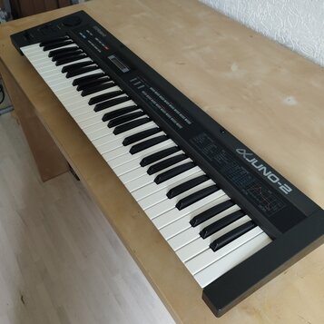 Černý elektronický klávesový nástroj s bílými a černými klávesami. Na pravé straně je bílý nápis JUNO-2 s logem před názvem a v horní části je logo R…