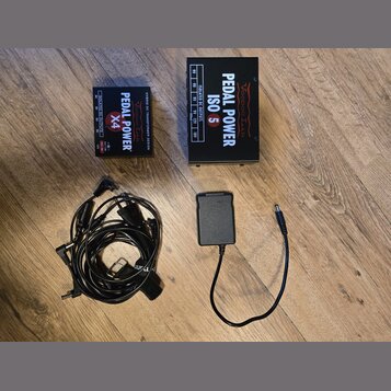 Na dřevěné podlaze jsou dvě černé napájecí zdroje pro efektové pedály a dva napájecí kabely. Menší zdroj má nápisy PEDAL POWER X4, ISOLATED DC OUTPUT…