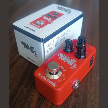Červený efekt pedál s nápisy tc electronic, HALL OF FAME 2 MINI a BYPASS. Na horní straně jsou tři černé oválové ovládací knoflíky a jeden stříbrný k…