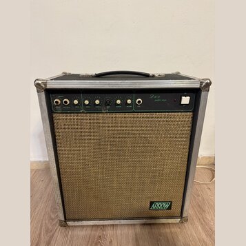 Kytarový kombo zesilovač v hliníkovém flightcase s černou rukojetí a kovovými rohy. Horní ovládací panel je zelený s bílými nápisy INPUTS, VOLUME, ov…