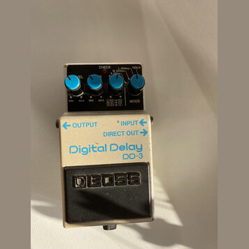 Kytarový efekt pedál značky BOSS, model Digital Delay DD-3. Má stříbrné kovové tělo s modrými ovládacími knoflíky a černým spínacím pedálem. Na horní…