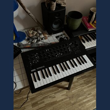 Černý syntetizátor s klaviaturou umístěný na podstavci. Na levé straně panelu je bílý nápis KORG minilogue xd a pod ním menší text POLYPHONIC HYBRID…