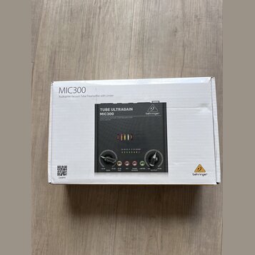 Fotografie znázorňuje bílý karton s produktem Behringer MIC300. Na krabici je nápis MIC300, Audiophile Vacuum Tube Preamplifier with Limiter a logo b…
