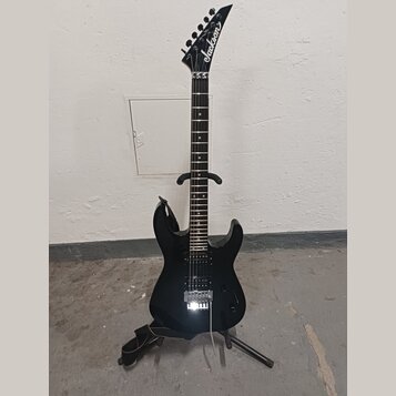 Černá elektrická kytara s špičatou hlavou, na které je bílé logo Jackson. Nástroj má tmavý hmatník a stříbrný kobylku typu Floyd Rose. Kytara je umís…