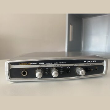 Stříbrné audio rozhraní s nápisy MOBILEPRE USB preamp | audio interface a M-AUDIO. Zařízení má dva regulační knofíky pro channel 1 a channel 2, každý…