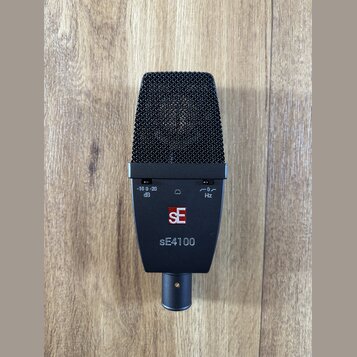 Černý dynamický mikrofon s kovovou mřížkou. Na těle jsou vidět přepínače s nápisy -10 0 -20 dB a 0 Hz, logo SE v červeném čtverci a modelové označení…