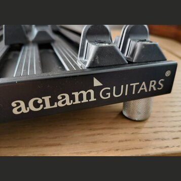 Detailní záběr na černý kovový most hudebního nástroje. Na boku je bílý nápis „aclam GUITARS“ s malým trojúhelníkem nad písmenem M a symbolem ® v pra…