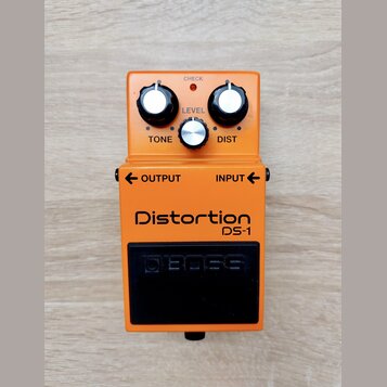 Oranžový kytarový efekt s nápisy Distortion a DS-1. Na horní části jsou tři ovládací knoflíky s popisky TONE, LEVEL a DIST oraz kontrolka CHECK. Ve s…