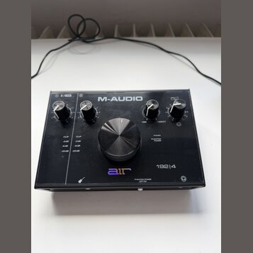 Černý audio rozhraní s nápisem M-AUDIO a označením modelu 192|4. Na předním panelu je velký centrální ovládací knoflík, čtyři menší knoflíky a logo a…