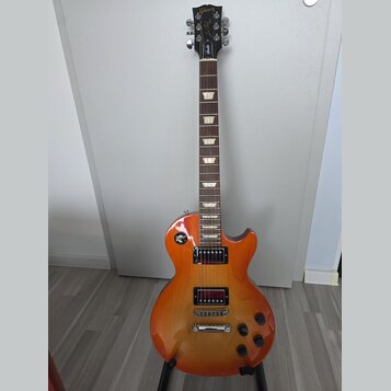 Elektrická kytara s trupem v žlutě-oranžovém sunburst provedení a hnědým hmatníkem s trapezoidními vložkami. Na hlavě je bílý nápis Gibson a kurzivní…