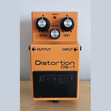 Oranžový kytarový efekt s nápisy Distortion a DS-1. Na horní části jsou tři ovládací knoflíky s popisky TONE, LEVEL a DIST a červená kontrolka CHECK.…