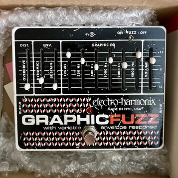 Kytarový efekt pedál v bílém rámu s černým ovládacím panelem. Na spodní části je nápis GRAPHIC FUZZ s přívlastkem with variable envelope response a l…