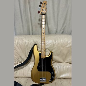 Zlatá elektrická basová kytara s černým pickguardem a javorovým hnedým hmatníkem. Na hlavě nástroje je nápis Fender Precision Bass. Kytara má čtyři s…