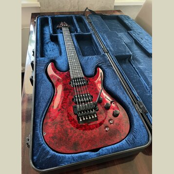 Elektrická kytara s červeným mramorovaným povrchem těla v černém tvrdém kufru s modrou výstuhou. Nástroj má šest strun, dva humbuckery a dva ovládací…
