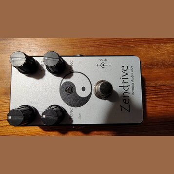 Stříbrný kytarový efekt s nápisem Zendrive a menším textem Hermida Audio USA. Na horní straně jsou čtyři černé otočné knoflíky s popisky Gain, Voice…