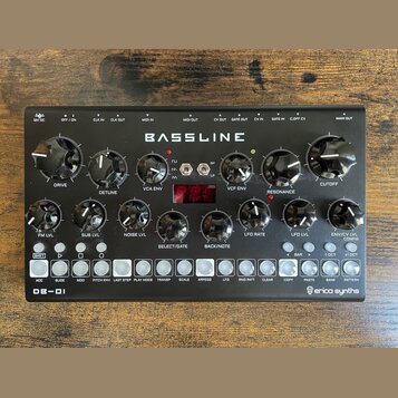 Černý syntezátor s nápisem BASSLINE a DB-01 v levém dolním rohu. V pravém dolním rohu je logo erica synths. Přístroj obsahuje několik černých otočnýc…