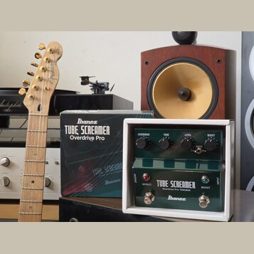 Na fotografii je zelený kytarový efekt v bílém ochranném boxu s nápisy TUBE SCREAMER, Overdrive Pro TS808DX a značkou Ibanez. Vedle efektu je umístěn…