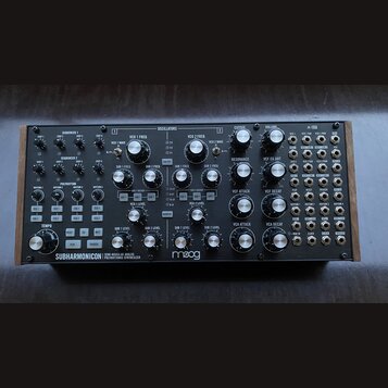 Černý semi-modulární analogový syntetizátor s dřevěnými bočními panely. Na spodní liště je bílý nápis SUBHARMONICON SEMI-MODULAR ANALOG POLYRHYTHMIC…