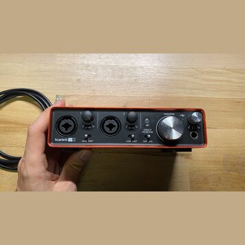 Červený audio rozhraní s černým předním panelem. Na zařízení jsou nápisy Scarlett 2i2, LINE INST, GAIN, DIRECT MONITOR, OFF ON, 48V a MONITOR. Jsou z…