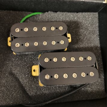 Dvě elektrické kytarové snímače typu humbucker s černými kryty. Snímače jsou umístěny na šedé pěnové výplasti v krabici. Na snímačích nejsou viditeln…