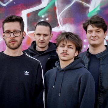 Na fotografii jsou čtyři muži v tmavém oblečení před barevnou zdí s graffiti. Jeden z nich má na černé mikině bílé logo adidas. Žádné hudební nástroj…