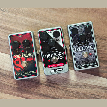 Tři efektové pedály značky electro-harmonix v kovových stříbrných šasi. První pedál je červený s nápisy Satisfaction, FUZZ a webovou adresou WWW.EHX.…