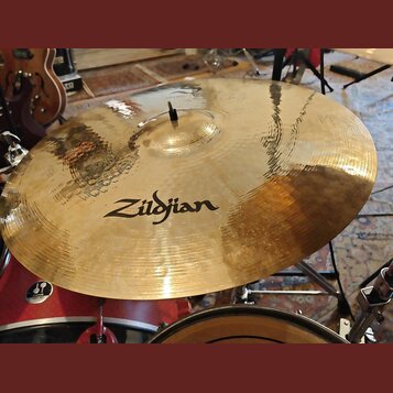 Zlatavý kovový činel s viditelným kovánívým povrchem a mírným opotřebením. Uprostřed je černý nápis Zildjian. Činel je osazen na stojanu v rámci bicí…