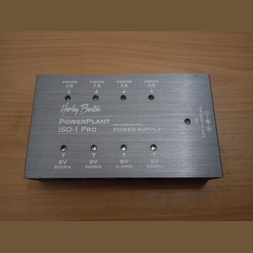 Stříbrný obdélníkový zdroj napájení z kovu s nápisy Harley Benton, PowerPlant ISO-1 Pro a ISOLATED OUTPUT POWER SUPPLY. Na horní straně jsou čtyři vý…